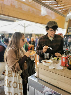 A man and a women standing at a street food market stand, eating small dishes from disposable plates.