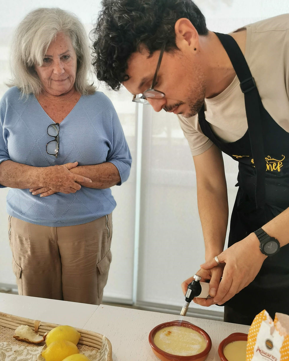 A Spanish chef wearing an apron and an elder woman are in a cooking class. He is burning the sugary coat of a typical Spanish dessert while she watches the method.