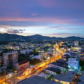 A vibrant cityscape at dusk with illuminated streets, buildings, and mountains in the background under a colorful, cloudy sky.