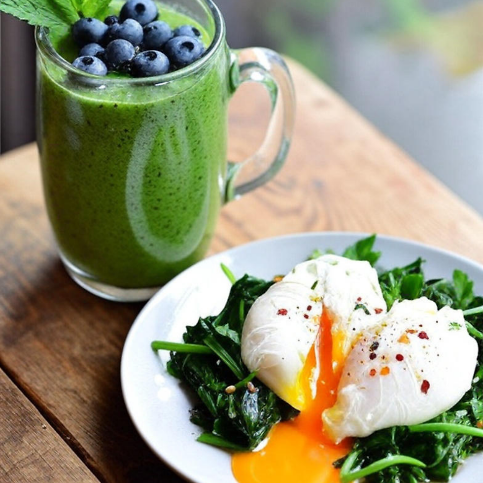 A green smoothie topped with blueberries and mint beside a white plate of poached eggs on spinach, with yolk oozing out, on a wooden table.