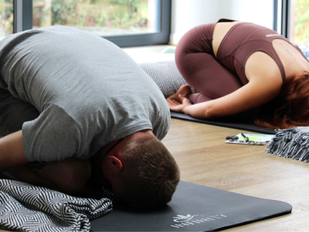 Two people in child’s pose on yoga mats indoors. One wears gray, the other maroon. A patterned blanket and cards nearby. Calm setting.