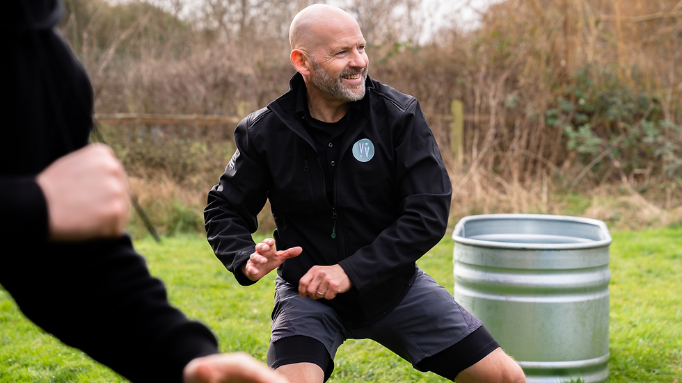 A man dressed in a black jacket in a squat stance practicing the Wim Hof Method, smiling, an ice bath in the background.