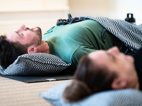 Two people lying down during a guided breathwork or relaxation session, resting with hands on their bodies beside a large window.