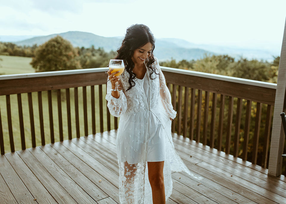 Bride getting ready on the porch of Cabin Heaven’s cabin, sipping mimosas and sharing makeup time with her wedding party at a Blue Ridge Mountain wedding venue.