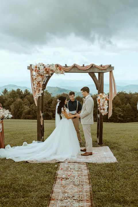 Bride and groom holding hands during their wedding ceremony at Cabin Heaven, standing on a boho rug aisle runner with mountain views in the background.