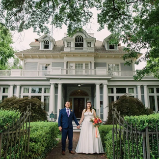 bride and groom pose in front of old style white house with hedges