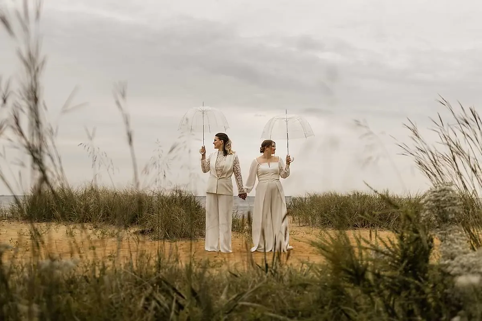brides hold hands and hold umbrellas while standing on a beach with bushes