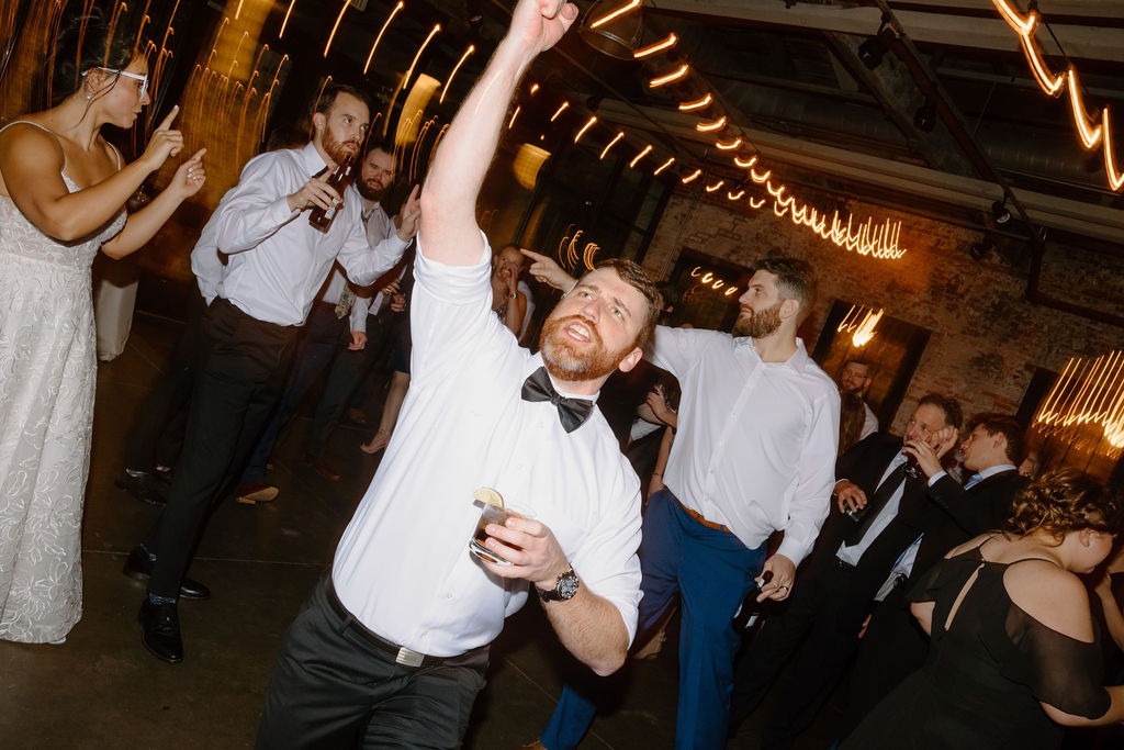 guy wearing black bow tie dancing with arm up and drink in the other hand while at wedding