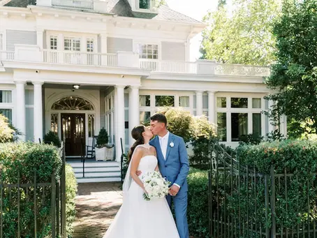 Bride in white gown and groom in blue suit kiss in front of a large white house with greenery. Romantic, sunlit setting.