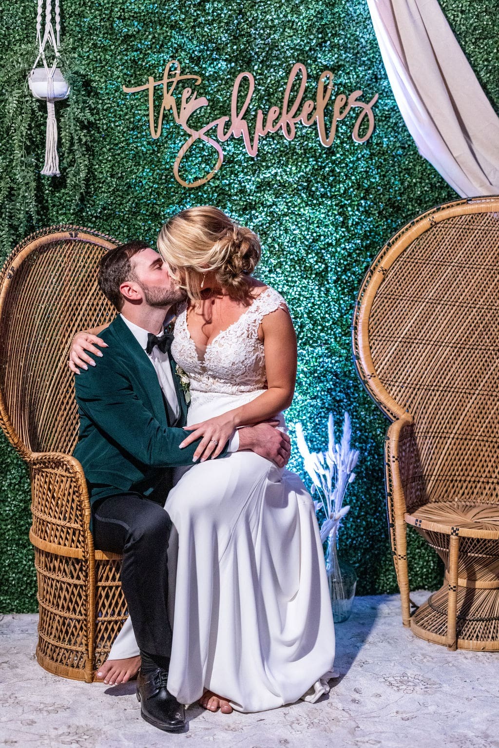 bride sitting in groom's lap in a wicker high-backed chair in front of green wall and kissing him