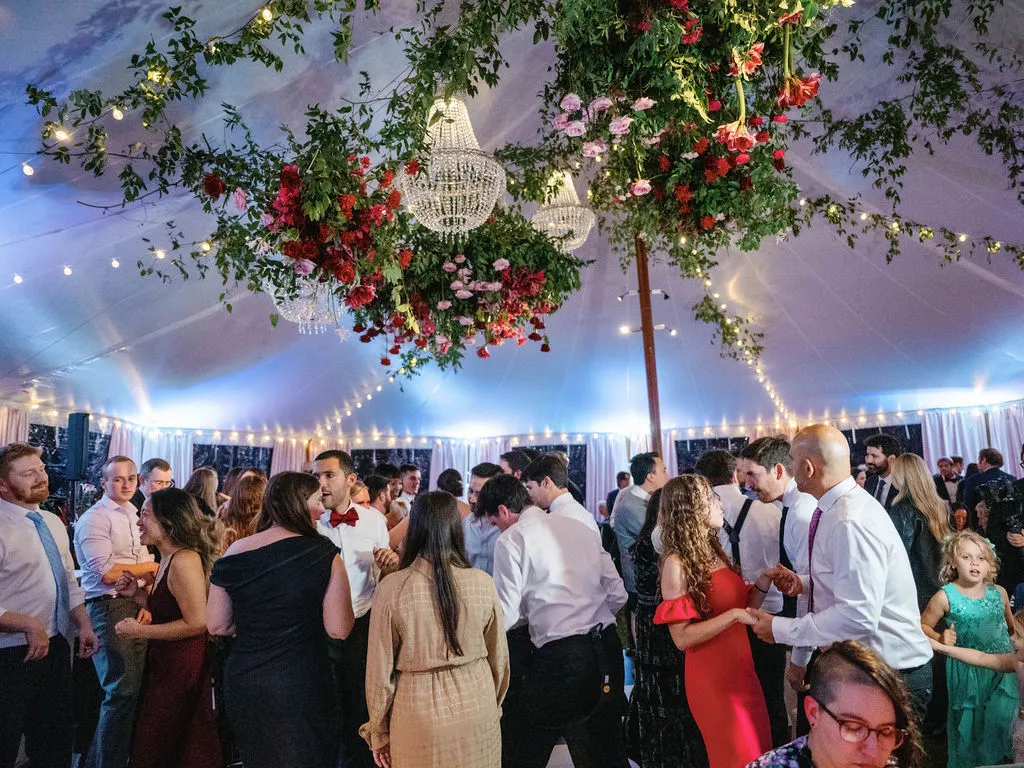 guests dancing in tent underneath floral and chandelier arrangement