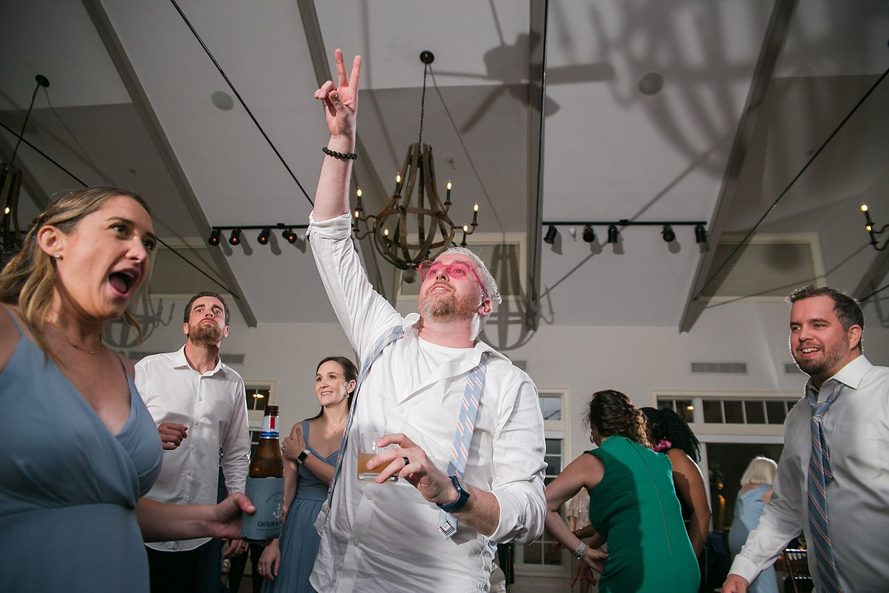 groom with pink glasses on dancing with hand up surrounded by guests at wedding