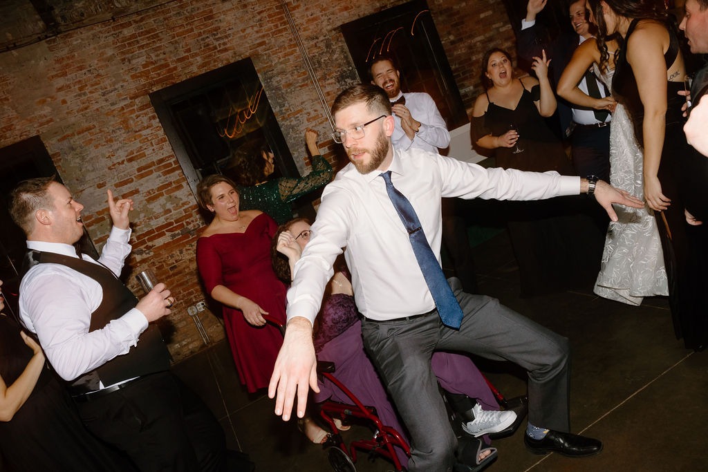 guy in blue tie dancing in front of woman in purple dress sitting on a mobile walker at wedding