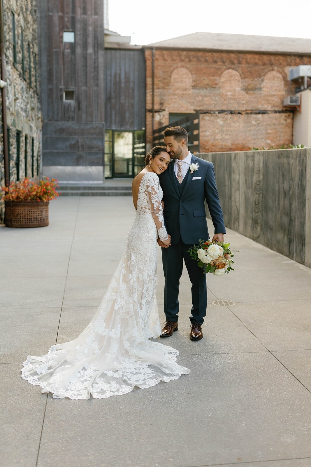 bride leans head on shoulder of groom while he holds bouquet while they stand outside of old industrial brick buildings