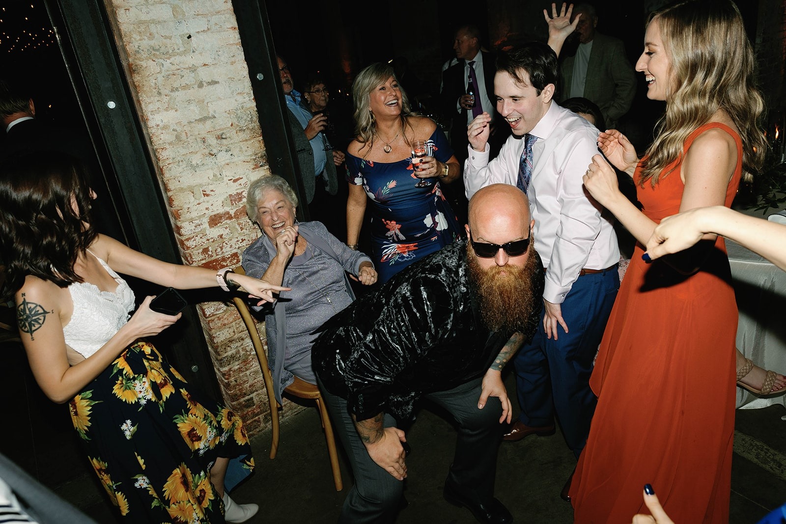 bald man with long beard wearing sun glasses shaking butt in front off smiling grandmother seated in a chair on dance floor at wedding