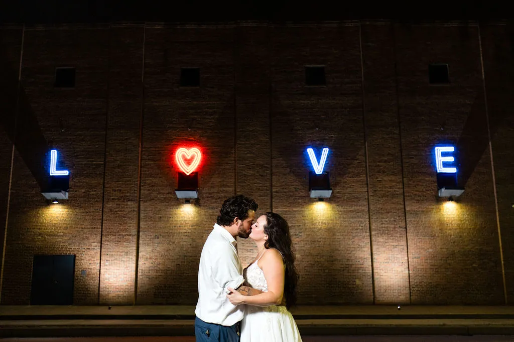 bride and groom kiss out front of LOVE neon sign on brick wall