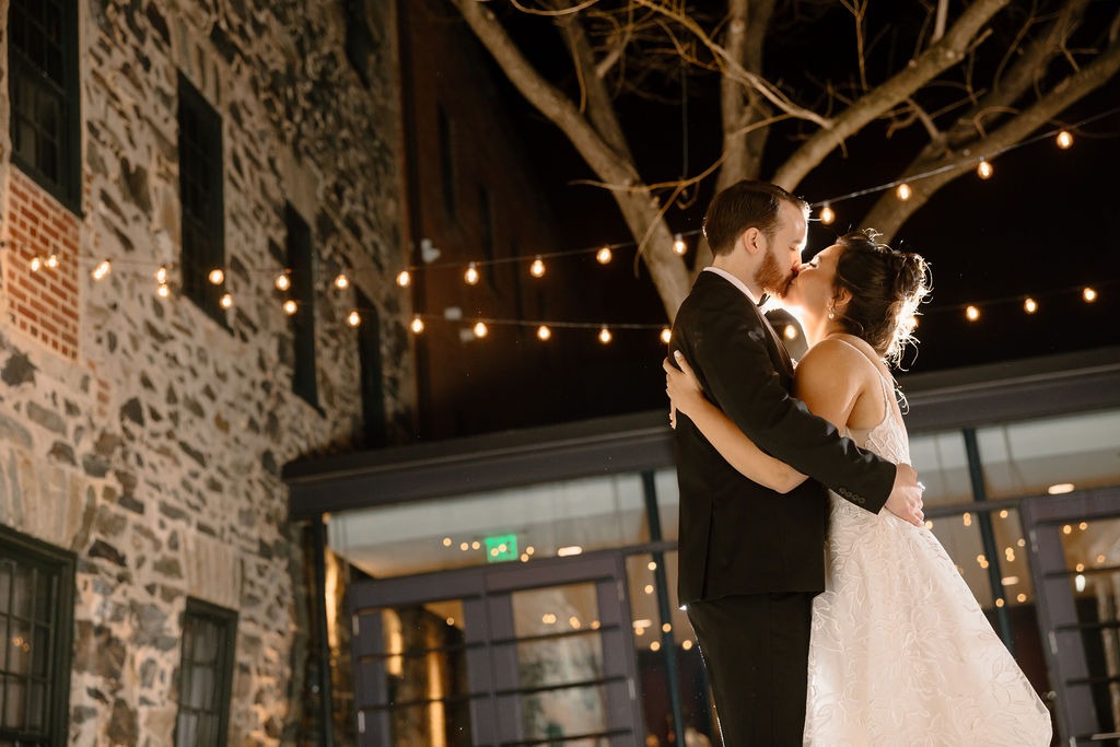 bride and groom kiss in courtyard of old industrial building with string lights hung up at night