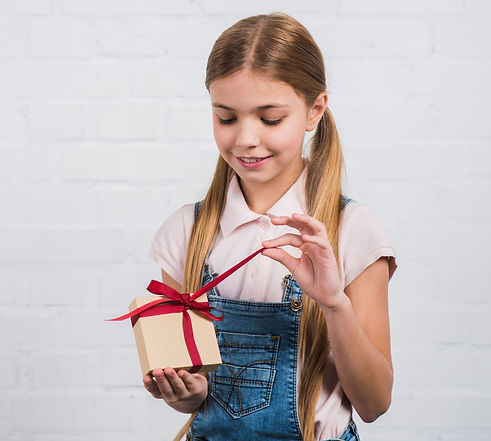 Young girl smiling as she opens a small gift box with a red ribbon, representing the joy of giving and receiving during the holiday season.