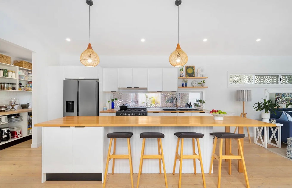 Modern white kitchen with pendant lights, a wooden island, stools, and a steel fridge. Open shelves display decor. Bright, airy mood.