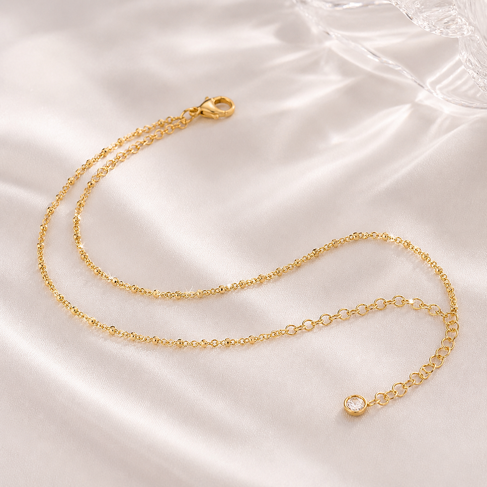 Delicate gold anklet with bead accents and crystal charm styled on silky neutral fabric.