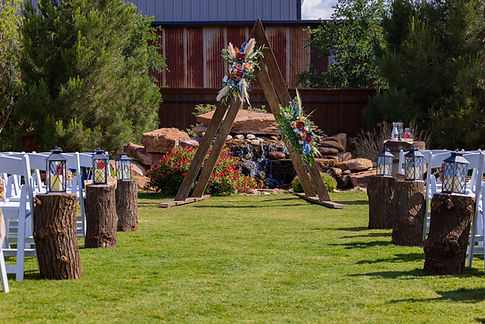 Outdoor wedding setting with wooden arch, floral arrangements, and lanterns on tree stumps.