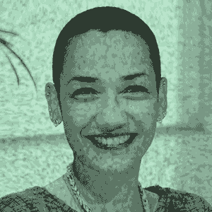 Uma mulher de pele preta não retinta, cabelos raspados, sorri com olhar para frente. A imagem está em tons de verde e preto e aparece com um efeito de televisão antiga, apresentando pequenos ruídos visuais.
