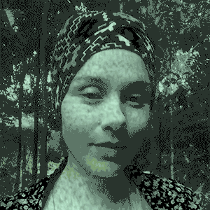 Uma mulher branca com olhos pequenos e castanhos e de turbante com desenhos dos símbolos adinkras nas cores vermelho, preto, azul e branco, tem o olhar direcionado para frente. A imagem está em tons de verde e preto e aparece com um efeito de televisão antiga , apresentando pequenos ruídos visuais.