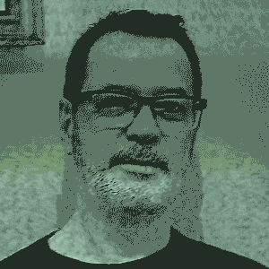 Um homem, de pele clara, cabelo curto e escuro, barba e bigode grisalhos, óculos de grau, vestindo camisa clara de gola, tem o olhar direcionado para frente. A imagem está em tons de verde e preto e aparece com um efeito de televisão antiga , apresentando pequenos ruídos visuais.