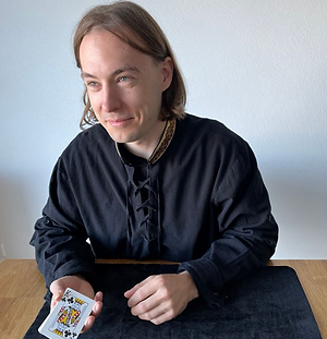 Zauberer Cardamon in schwarzem Gewand sitzt an einem Tisch und hält ein Kartenspiel in der Hand.