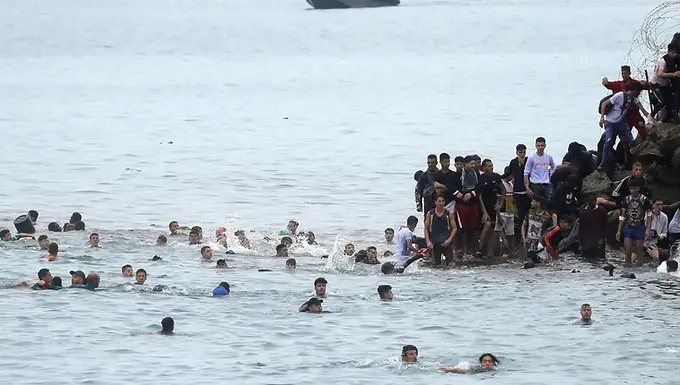 لقي 57 شخصاً على الأقل حتفهم خلال محاولة عبور إلى الجيب الإسباني سبتة، وذلك عقب حركة هجرة غير نظامية شملت عشرات الآلاف من المغاربة على الحدود بين إسبانيا والمغرب. وأفادت السلطات الإسبانية بأن جزءاً من الضحايا لقوا حتفهم غرقاً، فيما سُحق جزء آخر أثناء محاولتهم تسلق كاسر الأمواج الذي يدعم السياج الحدودي. وبينما كثّفت مدريد والرباط عمليات المراقبة وإعادة المهاجرين، أثارت الحادثة ردود فعل سياسية في عدة دول من الاتحاد الأوروبي. أعلنت السلطات الإسبانية، يوم الجمعة 31 تموز/يوليو 2026، أنها تمكنت من احتواء التدفق الهجري المسجّل منذ صباح الخميس باتجاه سبتة، الجيب الإسباني الواقع في شمال إفريقيا. ووفقاً لوزارة الداخلية الإسبانية، فإن نحو 50 ألف شخص عبروا الحدود براً وبحراً خلال تلك الفترة، كان نحو 48.300 منهم قد عادوا إلى المغرب بحلول الساعة السادسة مساء الجمعة (الساعة الرابعة عصراً بتوقيت غرينتش). وقال رئيس حكومة سبتة، خوان خيسوس فيفاس، إن عدد الأشخاص الذين عبروا الحدود خلال اليومين السابقين قد يصل إلى 60 ألفاً. أفاد ممثل الحكومة الإسبانية في سبتة بأنه تم انتشال 57 جثة من الجانب الإسباني للحدود، وحذّر من احتمال وجود ضحايا إضافيين في الأراضي المغربية. ووفقاً للسلطات، فإن بعض الأشخاص لقوا حتفهم غرقاً أثناء العبور البحري، بينما سُحق آخرون أثناء محاولتهم تجاوز كاسر الأمواج والهيكل المعدني الفاصل بين الإقليمين. استخدمت قوات الأمن المغربية الهراوات والغاز المسيل للدموع لمنع محاولات جديدة لدخول سبتة. وتمركزت فرق التدخل السريع عند مداخل الجيب الإسباني، في حين استُخدمت شاحنات مزودة بمدافع مائية لتفريق المجموعات التي ظلت متجمعة قرب السياج الحدودي. وأفادت وكالة رويترز برؤيتها حطام حافلة وسبع سيارات محترقة في المنطقة، نتيجة الاشتباكات التي وقعت خلال العملية. على الجانب الإسباني من الحدود، جرى توزيع آليات عسكرية على طول مقاطع مختلفة من السياج. أما في الأراضي المغربية، فقد بقي عشرات المهاجرين على التلال القريبة يراقبون التحركات، بينما بدأ مئات الأشخاص بالعودة عبر نقاط التفتيش أو عبر الفتحات الموجودة في الهيكل الفاصل بين البلدين. روى مهاجرون تحدثوا إلى رويترز معاناتهم في البقاء في سبتة بعد العبور. قال شاب مغربي من طنجة: "بصراحة، لا أعرف حتى لماذا جئت، والآن أنا عائد. لم آكل منذ غداء أمس، رغم أنني أحضرت معي بعض المال... ما نفعله ليس جيداً ولا ممتعاً". أما مهاجر آخر، عُرف فقط باسم أيمن، وهو حلاق يبلغ من العمر 20 عاماً من مدينة العرائش، فقال لرويترز إنه سبح نحو خمس ساعات للوصول إلى سبتة. ووفقاً لروايته، "لم يكن هناك طعام في سبتة، وقد طُردنا بقسوة من قبل الجيش الإسباني". زار رئيس الوزراء الإسباني بيدرو سانشيز سبتة يوم الجمعة، واستُقبل بمظاهرات احتجاجية. وخلال الزيارة، وصف الدخول الجماعي بأنه "انتهاك للسيادة الترابية لإسبانيا"، وقال إن الحكومة تسرّع عملية إعادة من دخلوا بطريقة غير نظامية، بالتنسيق مع السلطات المغربية. سبتة ومليلية هما المدينتان الإسبانيتان المستقلتان الواقعتان في شمال إفريقيا، وتشكّلان الحدود البرية الوحيدة بين الاتحاد الأوروبي والقارة الإفريقية. وتشهد المدينتان محاولات عبور متكررة من قبل مهاجرين يسعون للوصول إلى الأراضي الأوروبية، رغم أن السلطات الإسبانية وثّقت التحرك الذي جرى بين الخميس والجمعة باعتباره واحداً من أكبر التحركات التي شهدتها المنطقة. أحدثت الحادثة تداعيات بين الحكومات الأوروبية. أعلنت إيطاليا تعليق تطبيق اتفاقية شنغن تجاه إسبانيا بالنسبة للسفر جوّاً وبحراً، وهو إجراء اعترضت عليه مدريد، التي قالت إنها تعتبر القرار غير متوافق مع معاهدات الاتحاد الأوروبي. أرجعت أحزاب يمينية في عدة دول أوروبية ازدياد عمليات العبور إلى السياسة الهجرية التي تبنّتها الحكومة الإسبانية، بما في ذلك تسوية أوضاع مئات الآلاف من المهاجرين التي نُفّذت خلال العام. ورداً على ذلك، استدعت الحكومة الإسبانية سفير إيطاليا للاحتجاج على تصريحات وزير الخارجية الإيطالي أنطونيو تاجاني، الذي قال إن سياسة العفو الإسبانية شجّعت على الاتجار بالبشر. علق الرئيس الأميركي دونالد ترامب على الحادثة خلال اجتماع للحكومة. ووفقاً له، فإن عمليات العبور في سبتة "تبدو كغزو"، وأضاف: "سيحدث لنا الشيء نفسه إذا لم يُنتخب الجمهوريون، ولكن بشكل أسوأ"، في إشارة إلى الانتخابات التشريعية النصفية. في فرنسا، أعلن وزير الداخلية لوران نونييز أن عدد عناصر الشرطة على الحدود مع إسبانيا سيُضاعف خمس مرات بحلول السبت، مع تعزيز المراقبة الجوية والدوريات السككية. ورغم أن منطقة شنغن تنص على حرية التنقل بين الدول المشاركة، فإن تشريعاتها تسمح بإعادة فرض الضوابط الحدودية مؤقتاً في حالات تُعتبر استثنائية. في منشور على منصة التواصل الاجتماعي إكس، قالت الجمعية الموحدة للحرس المدني (AUGC) إن عدد العناصر المكلفين بمراقبة سياج سبتة كان غير كافٍ في مواجهة الارتفاع المفاجئ في عدد الأشخاص المسجّل يوم الخميس، مما جعل السيطرة على الوضع صعبة، وفقاً للجمعية. صرّح الوزير الإسباني للسياسة الترابية، أنخيل فيكتور توريس، بأن أحد العوامل المرتبطة بزيادة محاولات العبور قد يكون قراراً صادراً مؤخراً عن المحكمة العليا الإسبانية يمنع الرفض الفوري للمهاجرين الذين يتم اعتراضهم في البحر أثناء محاولتهم الوصول إلى سبتة أو مليلية. غير أن القرار لا يمنع إجراءات الطرد من التنفيذ لاحقاً من قبل السلطات المختصة. على الجانب المغربي من الحدود، بقي آلاف المهاجرين في مدينة الفنيدق طوال الليل، رغم تعزيز القوات الأمنية. وعلى مدار يوم الجمعة، غادر جزء منهم المنطقة، بينما واصل آخرون التحرك على طول الشريط الساحلي بحثاً عن طرق بديلة للوصول إلى سبتة. وقال رجل عُرف فقط باسم إبراهيم، يبلغ من العمر 32 عاماً ويقيم في طنجة، إنه وصل إلى المكان بعد إغلاق المداخل، ووجد المعبر مغلقاً عملياً.