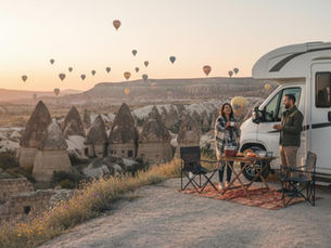 A couple enjoying breakfast outside their rented campervan during a sunrise in Cappadocia, Turkey, with hot air balloons in the background.
