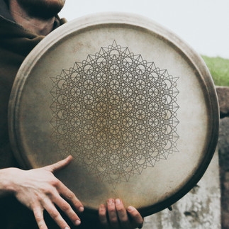 A traditional frame drum being held by a person. What makes this drum unique is the intricate sacred geometry pattern etched or printed on the drumhead surface. The design covering the drumhead is a radial geometric grid composed of repeating, interlocking circular motifs. The structure resembles isotropic triangle grids, tiling patterns similar to Islamic geometric art, and Flower of Life derivatives (expanded, layered hexagonal structures) Core components are a 12-fold symmetry, the star polygons and rosettes, the hexagonal tiling, and the central axis with radial uniformity. This kind of design is often believed to represent harmony, balance, and the interconnectedness of all life — perfect for an instrument associated with vibration and energy. Such geometric patterns have a cultural/spiritual significance used in meditative instruments for sound healing, visionary art and consciousness-expanding visuals, and representations of cosmic order and universal frequency.