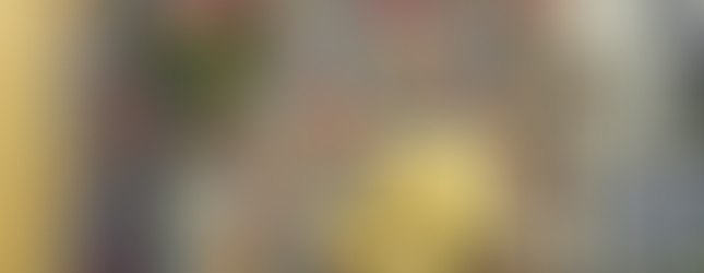 Light filters through sheer curtains, creating a cozy atmosphere in a Sims 4 room with a wooden floor, a yellow chair, some plants, all in a floral wallpapered room. Wallpaper cc by CarolaSims.