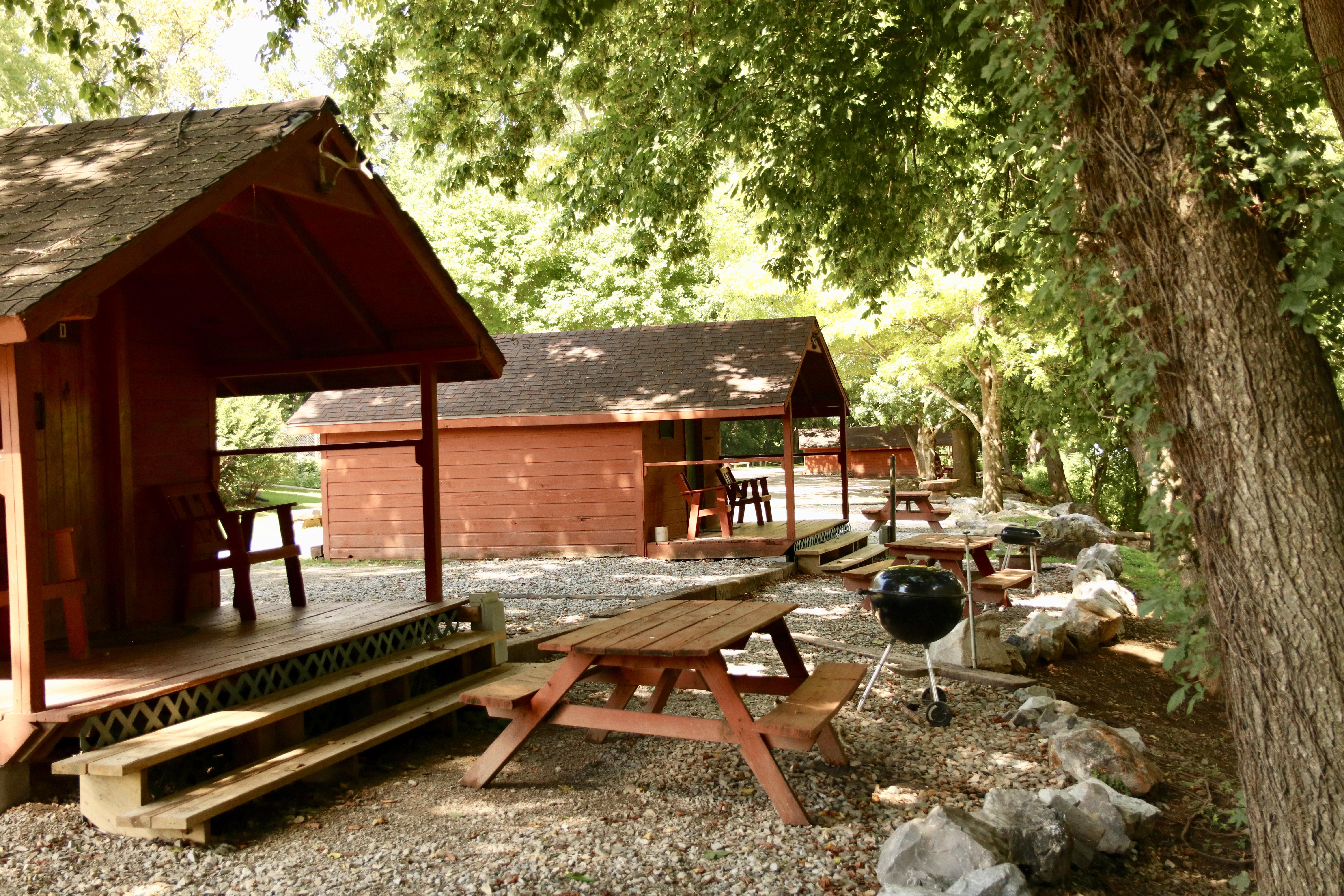 A wooden cabin with a deck and a picnic table in front of it, surrounded by trees and rocks.