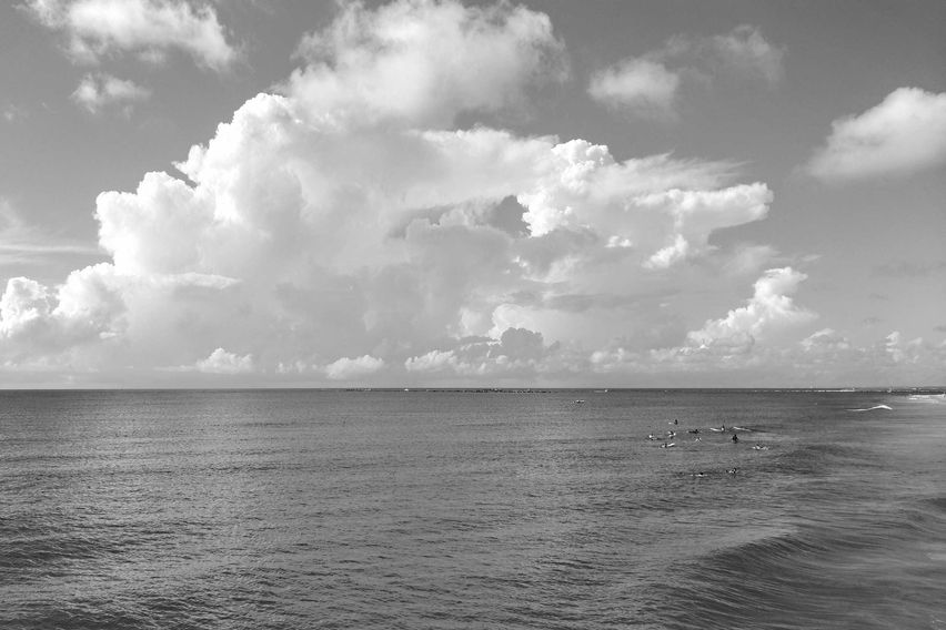 Black and white photo of the Atlantic Ocean and sky on a sunny day, with fluffy clouds, shimmering water, and tiny figures of board surfers in the distance.