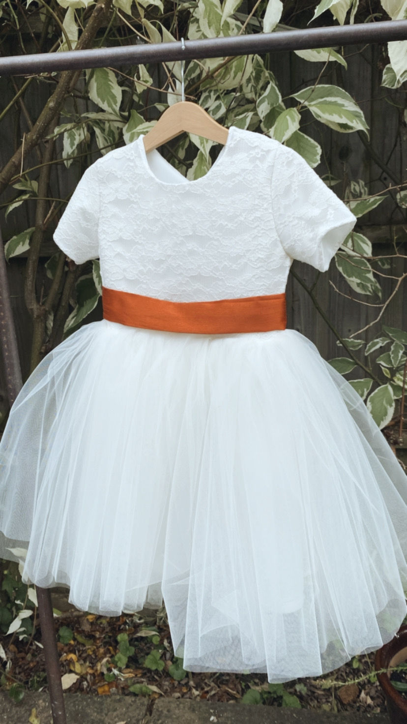 Thumbnail: A handmade ivory white lace cap sleeve dress with a V pearl button back and a soft tulle knee length skirt flower girl dress. It features a large burnt orange duchess satin bow sash.