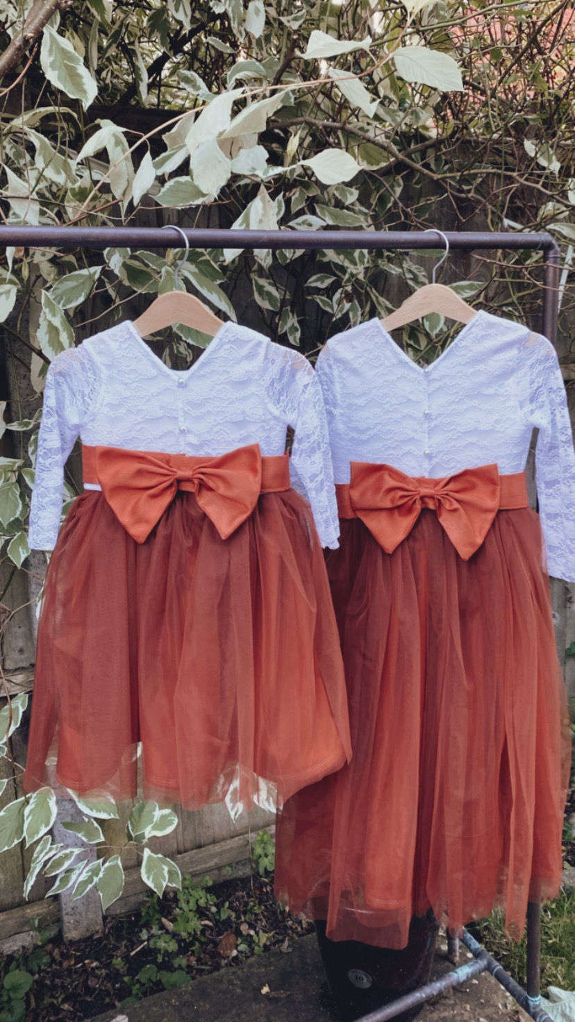 Thumbnail: A handmade ivory white lace top with sheer long sleeve lace sleeves a V pearl button back and has soft ankle length burnt orange tulle skirt. It features a large burnt orange duchess satin bow sash.