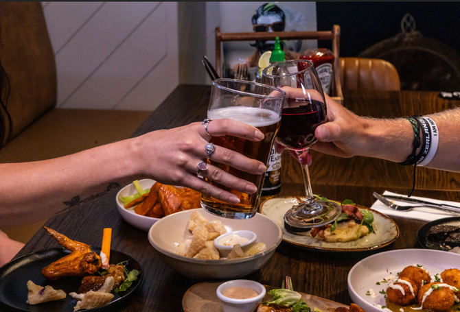 Couple sharing a toast with beer and wine alongside a selection of nibbles during a cozy date night at John Barleycorn’s Taphouse Newmarket, a top rooftop pub and dining spot in Newmarket Auckland.