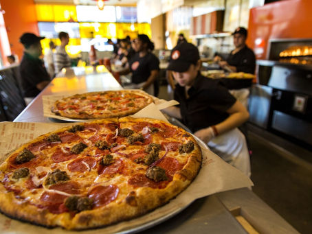 Interior of a Blaze Pizza restaurant showing staff in black uniforms preparing pizzas with pepperoni and sausage behind a counter under warm orange lighting.