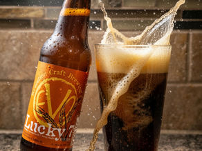 Beer bottle spilling dark lager into a foamy glass on a granite counter. Label reads "Lucky 7." Splash and droplets in motion.