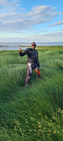 A colored man in motorbike wear in a grass area with lake in the background