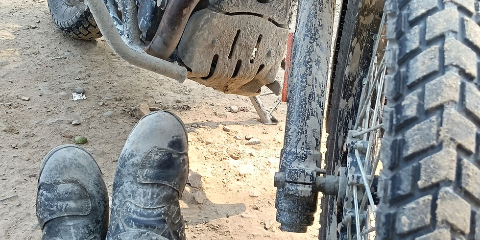 Dusty boots rest on a dirt road beside a muddy motorcycle tire and engine. Sunlit, rugged terrain.