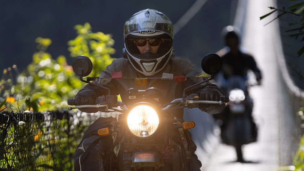 Motorcyclist in helmet rides on a bridge, headlights on. Bright foliage in background, secondary rider follows. Vibrant, adventurous mood.