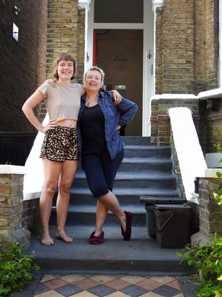 A caucasian mother ad daughter stand on the steps of their home, the daughter is wearing shorts and a T-shirt and the mother is wearing black leggings and a black top and a denim shirt.  It was a doorstep portrait photograph taken during the coronavirus lockdown in London 2020.  It is a coronavirus lockdown covid-19 doorstep portrait from empowerment photographer HeardinLondon.