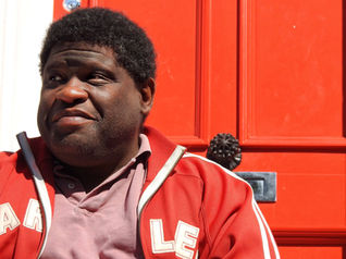 A portrait of a black man with short hair looking to the left.  He is wearing a red Addidas tracksuit top and a pink shirt and sits in front of a red door.  It is a landscape photograph.  It is a coronavirus lockdown covid-19 doorstep portrait from empowerment photographer HeardinLondon.