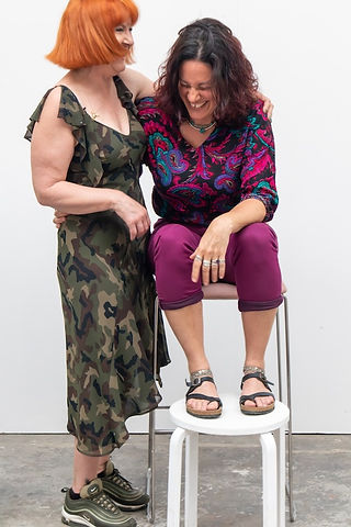 Two woman laugh.  On the left is a woman with orange hair and a green dress, standing.  A dual heritage woman wearing purple trousers and a red and green and purple shirt sits in front of a white background.  It is a portrait photograph taken by body confidence photographer HeardinLondon