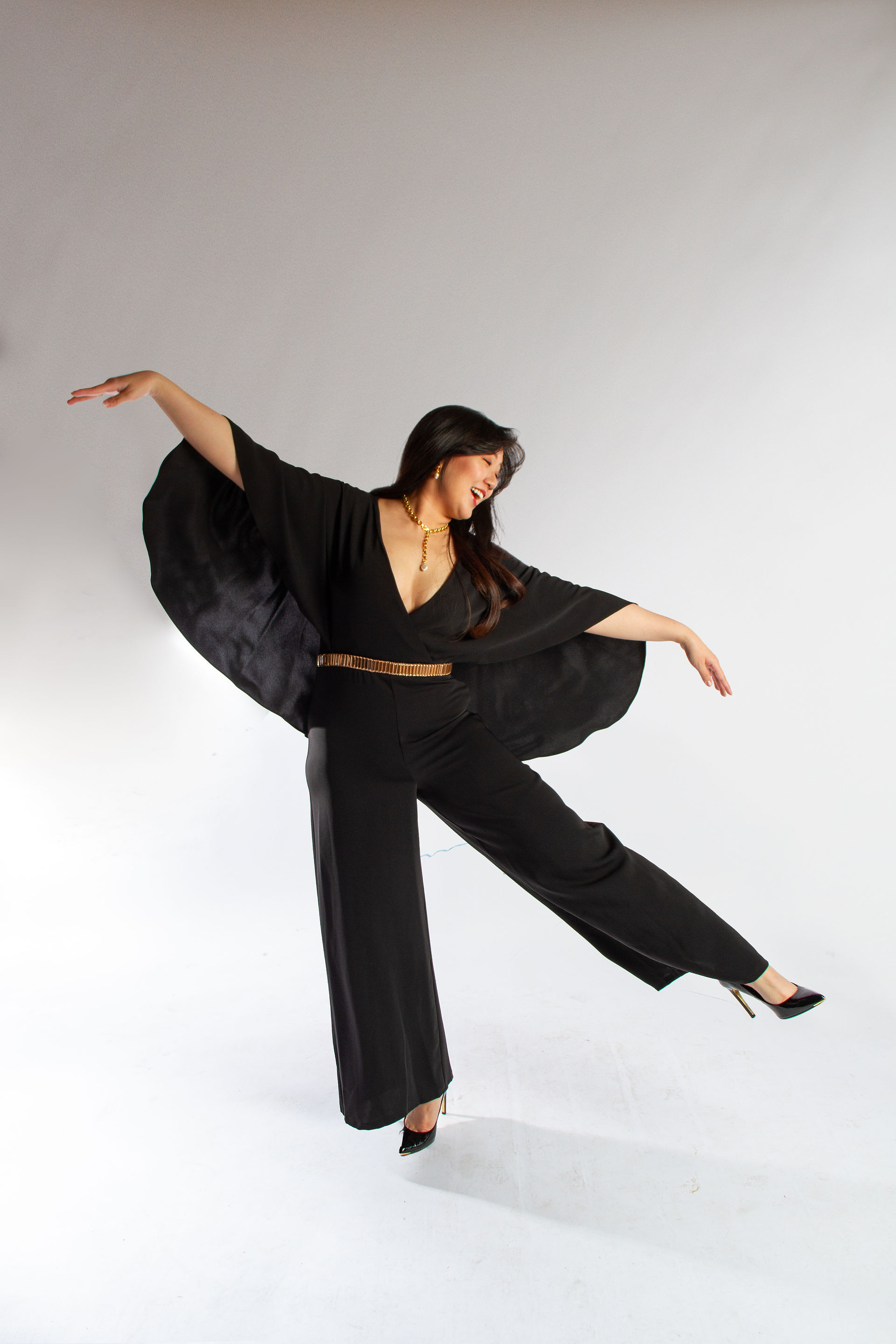 A Chinese woman in a black jumpsuit with a cape, dances.  It is a portrait photograph.