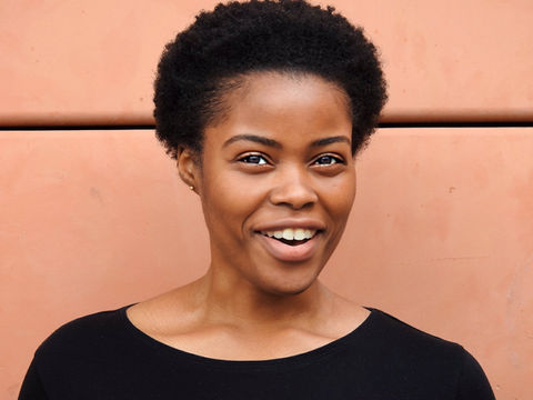 A black woman with short hair and a black long sleeve top stangs in front of a pink wall.  It is a doorstep portrait photograph taken during the coronavirus pandemic lockdown in London.  It is a coronavirus lockdown covid-19 doorstep portrait from empowerment photographer HeardinLondon.