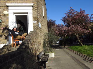 A caucasian man sits on a wooden chair in the doorway to his house. It is a doorstep portrait photograph taken during the coronavirus pandemic lockdown in London.  It is a coronavirus lockdown covid-19 doorstep portrait from empowerment photographer HeardinLondon.