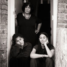 An Indian heritage woman stands in a hallway corridor behind her two daughters who are sat on the step of their house.   It is a doorstep portrait photograph taken during the coronavirus pandemic lockdown in London.  It is a coronavirus lockdown covid-19 doorstep portrait from empowerment photographer HeardinLondon.
